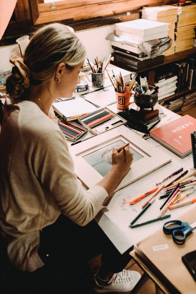 From above of young concentrated woman creating picture at table with many art supplies